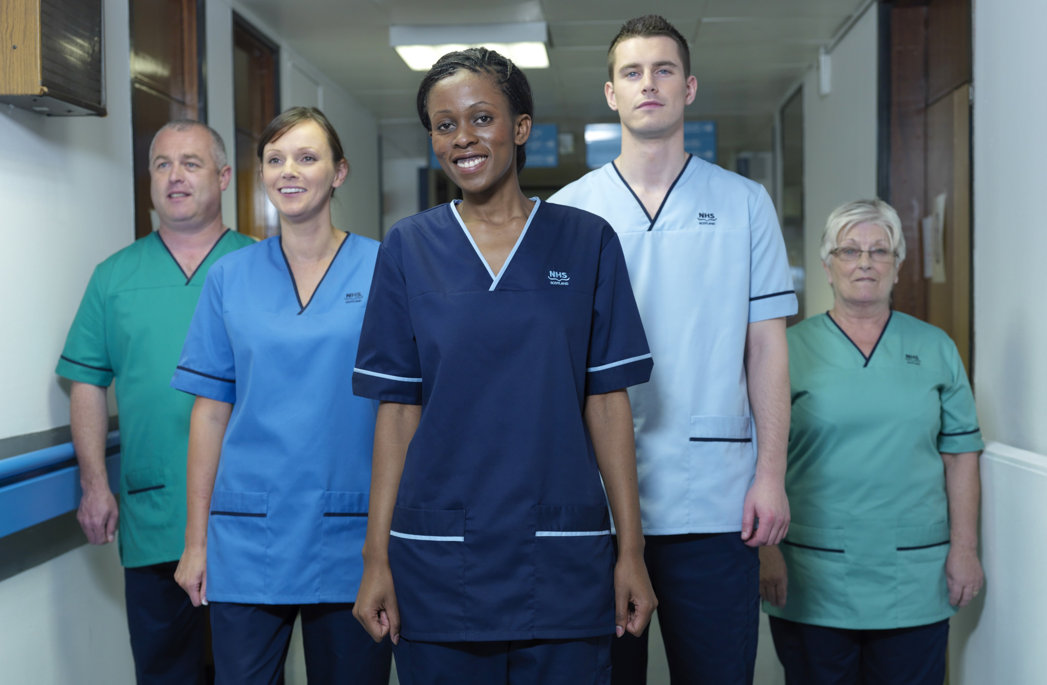 A group of NHS Scotland staff, including midwives, nurses and porters with the appropriate green and blue scrubs. They are standing in a line in a hospital corridor, looking towards the camera and smiling.