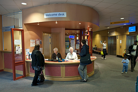 The welcome desk of an NHS Scotland hospital facility, with two receptionists sat behind it and people in the foyer.