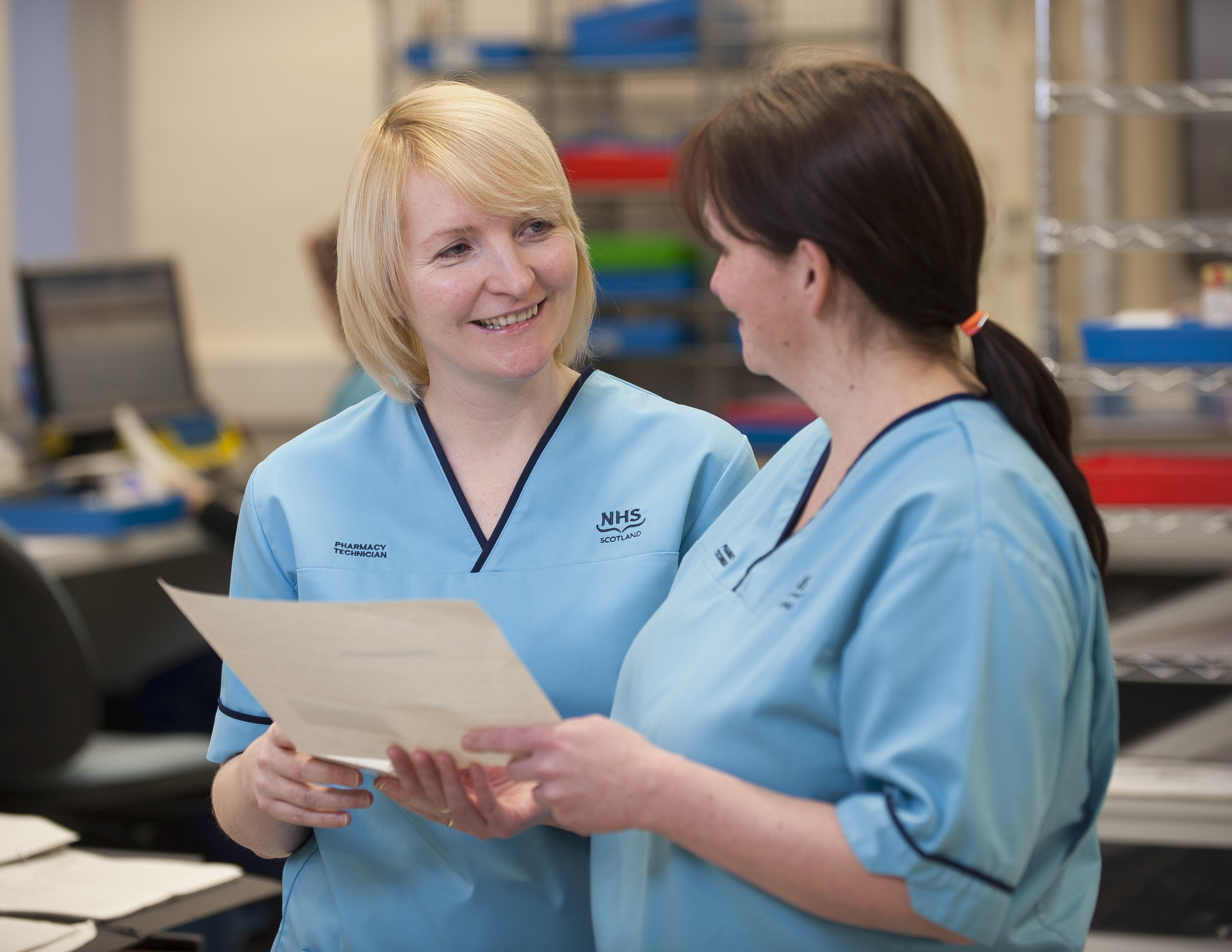 Two NHS Scotland nursing staff speaking to each other, smiling, at a nurses station.