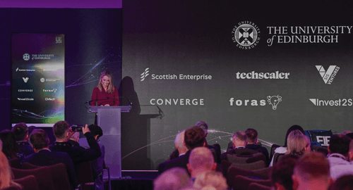 A still from the 2024 EIE conference, with a woman behind a lectern on stage, smiling in a red shirt. The backdrop is a University of Edinburgh banner.