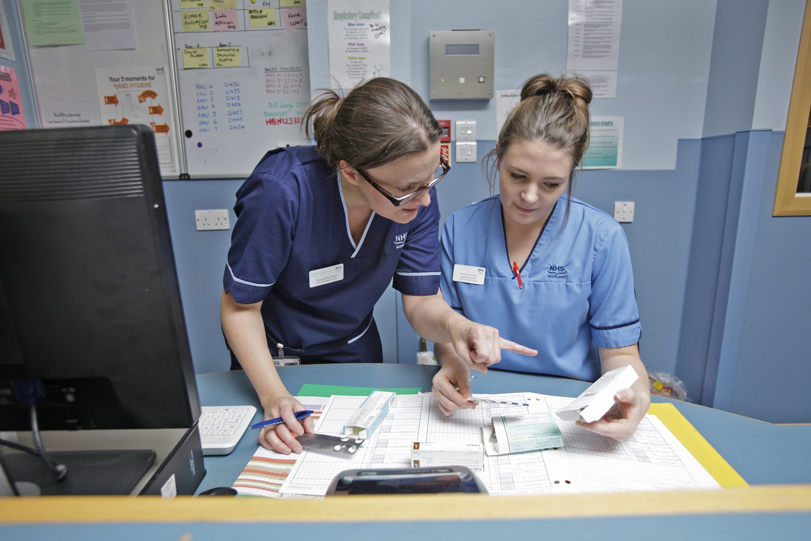 Two NHS Scotland nursing staff at a station in a hospital environment.