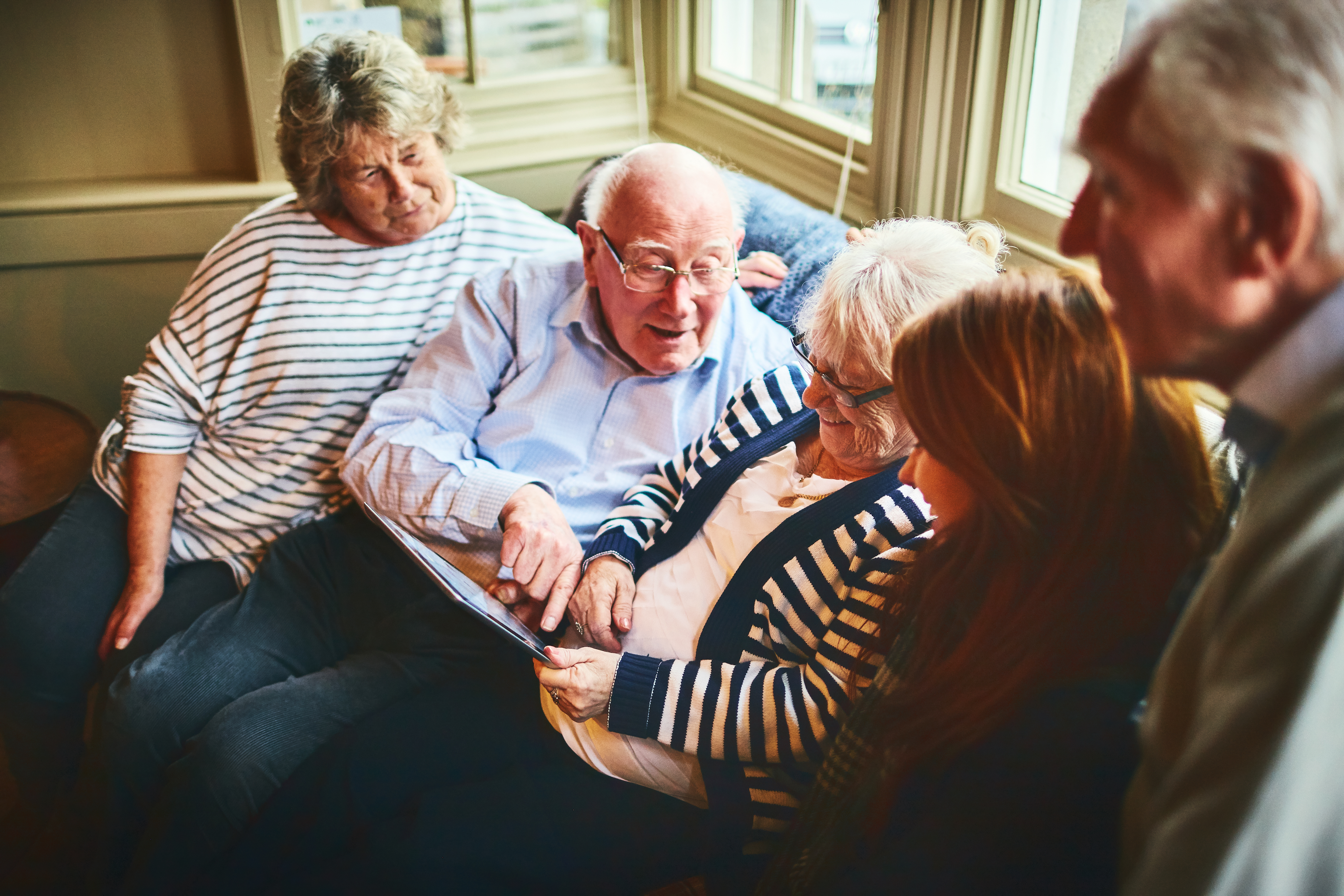 A group of older people gathered around a tablet, smiling and speaking to one another.