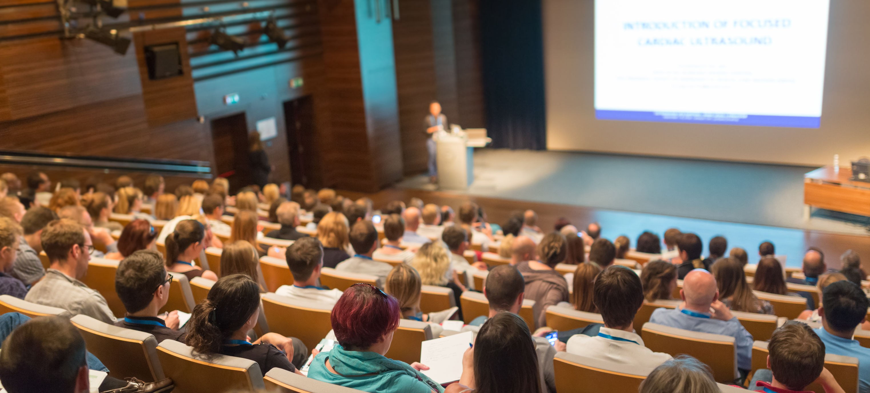 A conference hall, the photo looking down from the top of the busy lecture theatre towards the stage with a presenter.