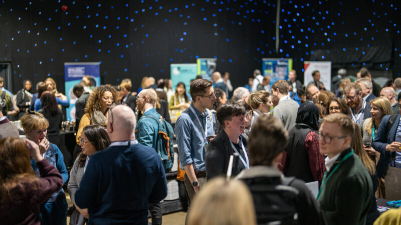 Picture of a previous HDR-UK conference, with a crowd of people networking in an exhibition hall surrounded by promotional graphics and stands.