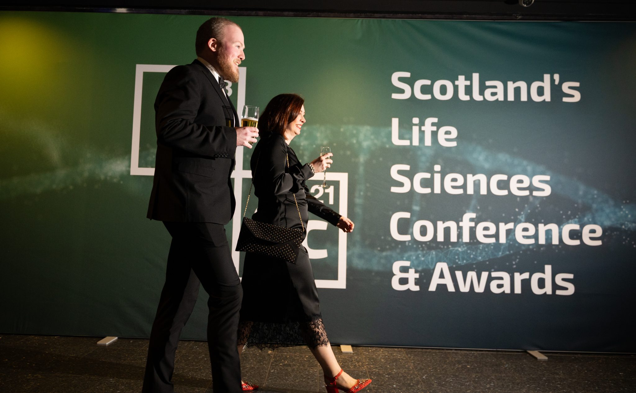 A couple in formal dress walking past a hoarding which reads, "Scotland's Life Sciences Conference and Awards".