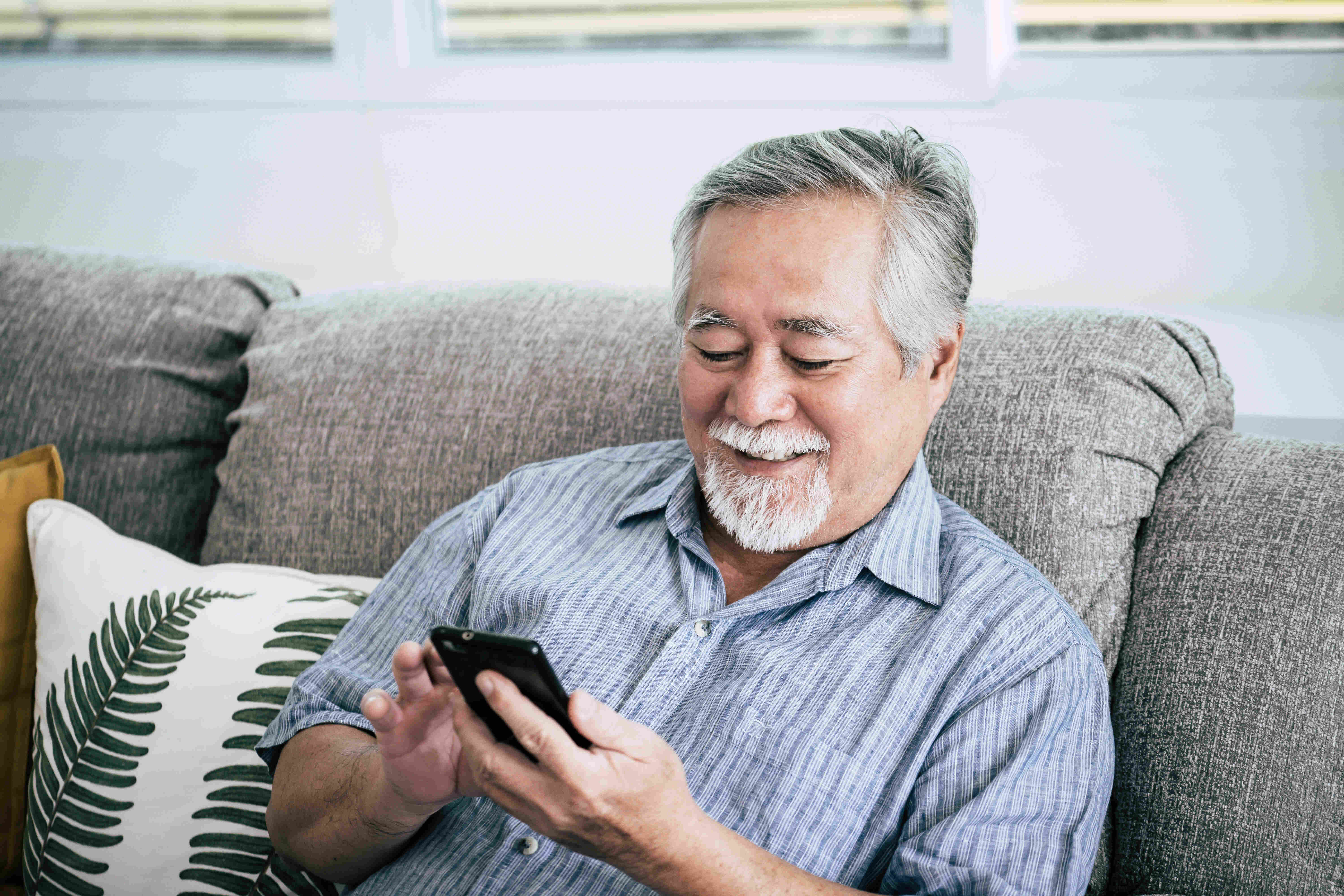 A senior man using a phone while sitting on a grey couch.