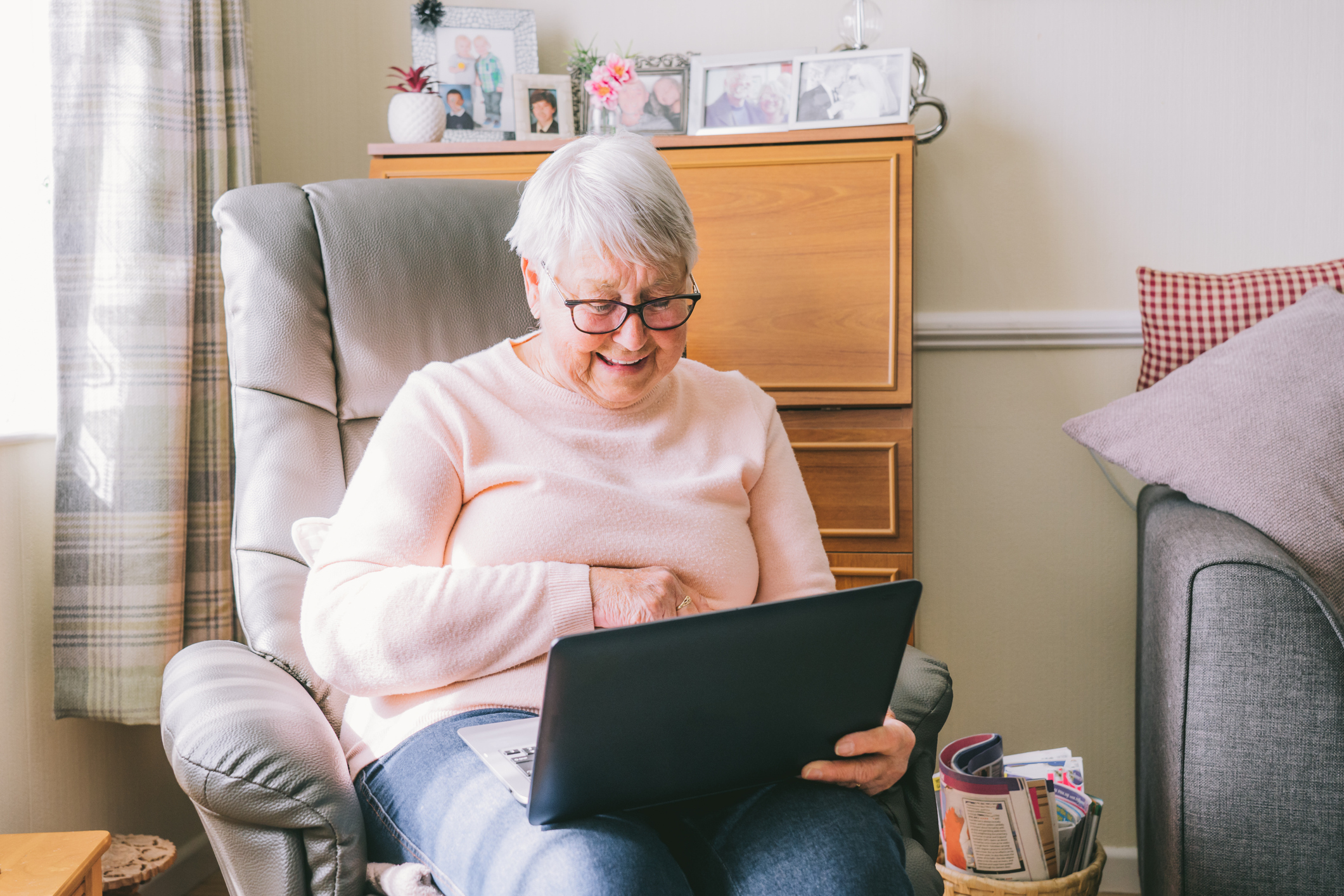 An older woman with glasses in an armchair at home, using a laptop and smiling broadly.