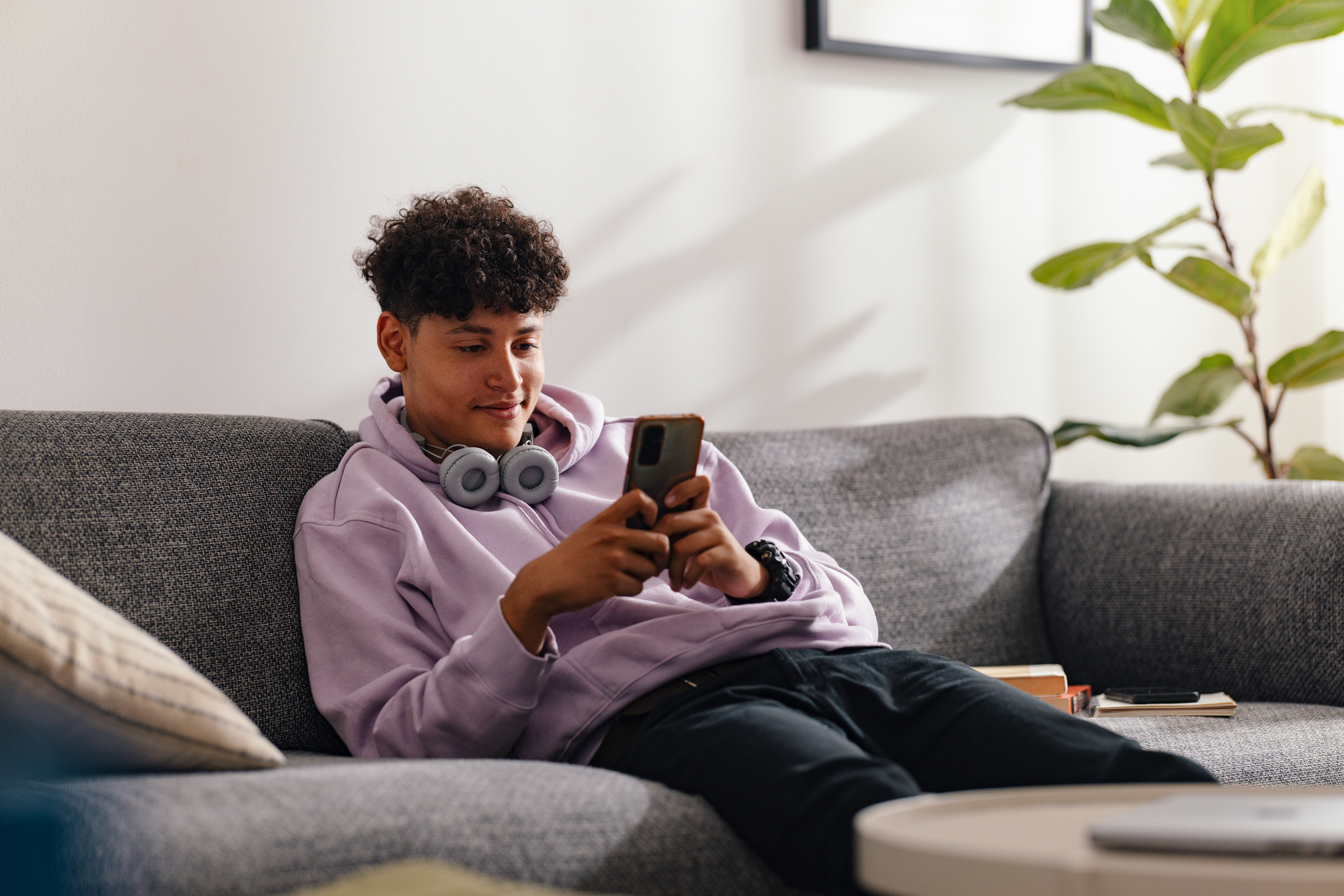 A teenage boy sitting on a couch, wearing headphones and using a smartphone.