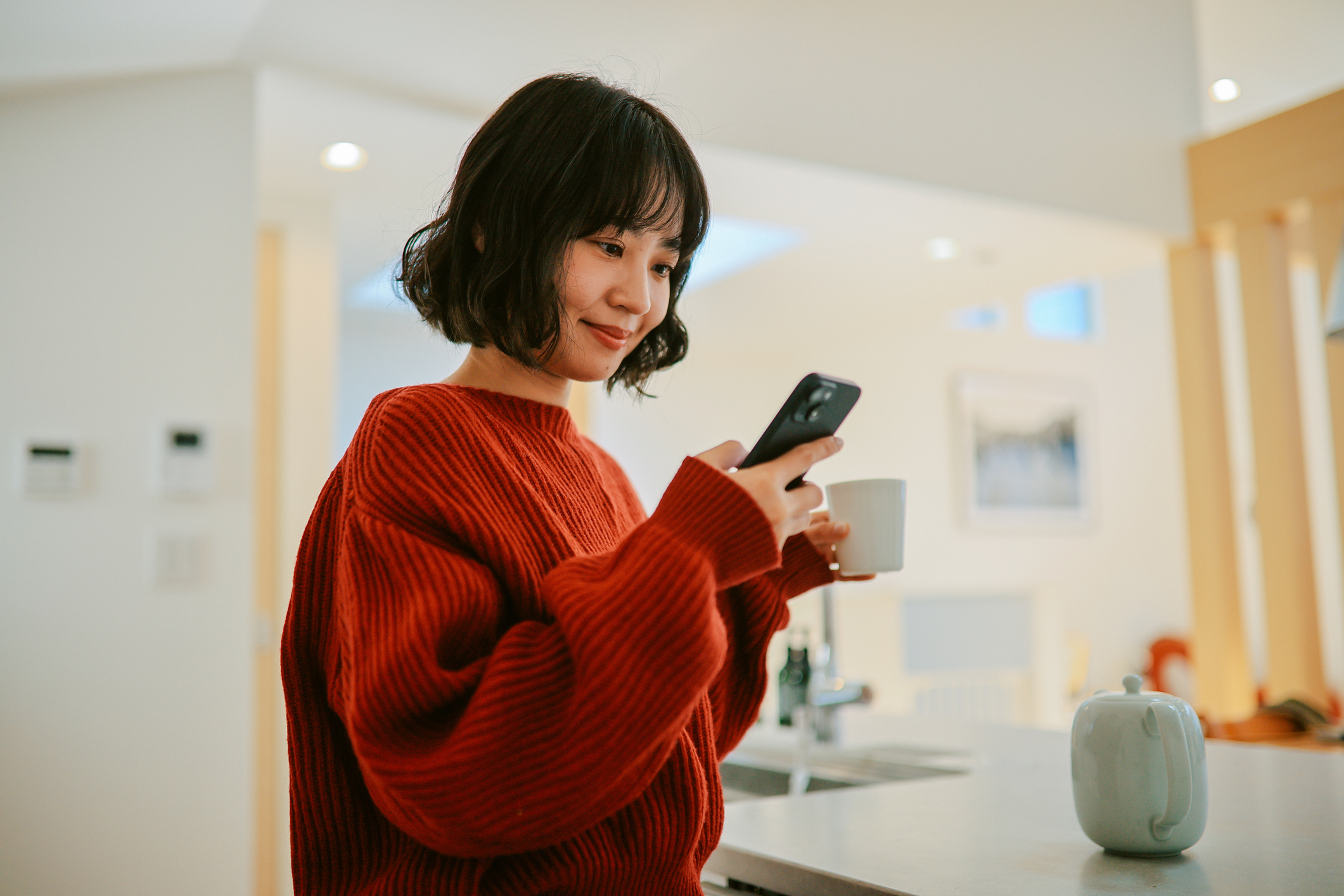 A woman looking at her phone in a kitchen. She has black hair in a bob and is wearing an orange jumper.
