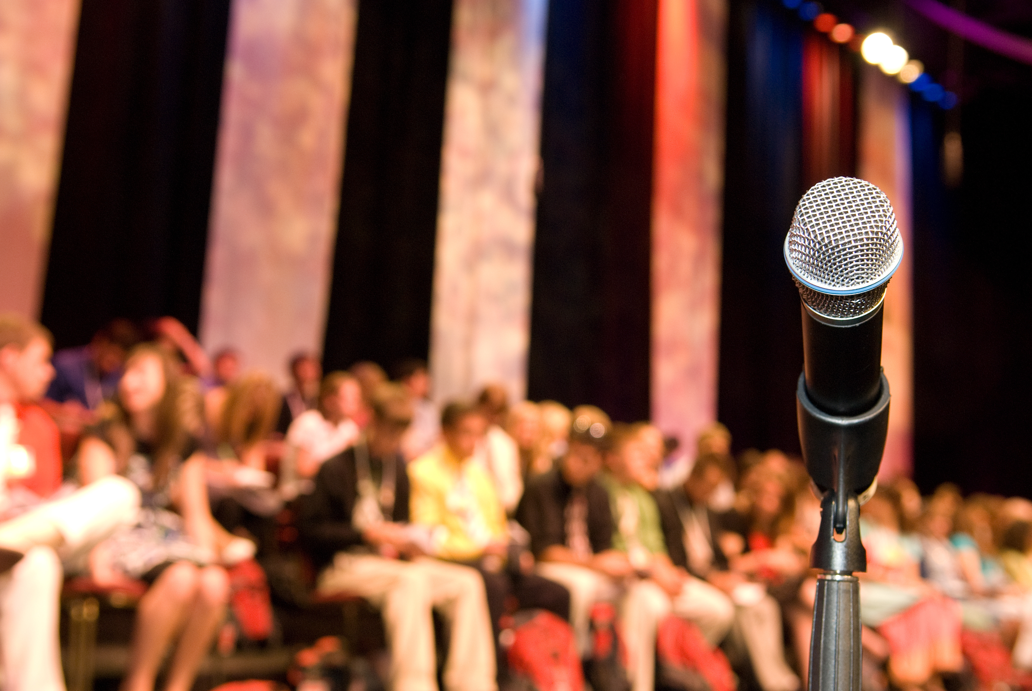 A conference photo with a microphone foregrounded, and a crowd waiting for a session to start in the background.