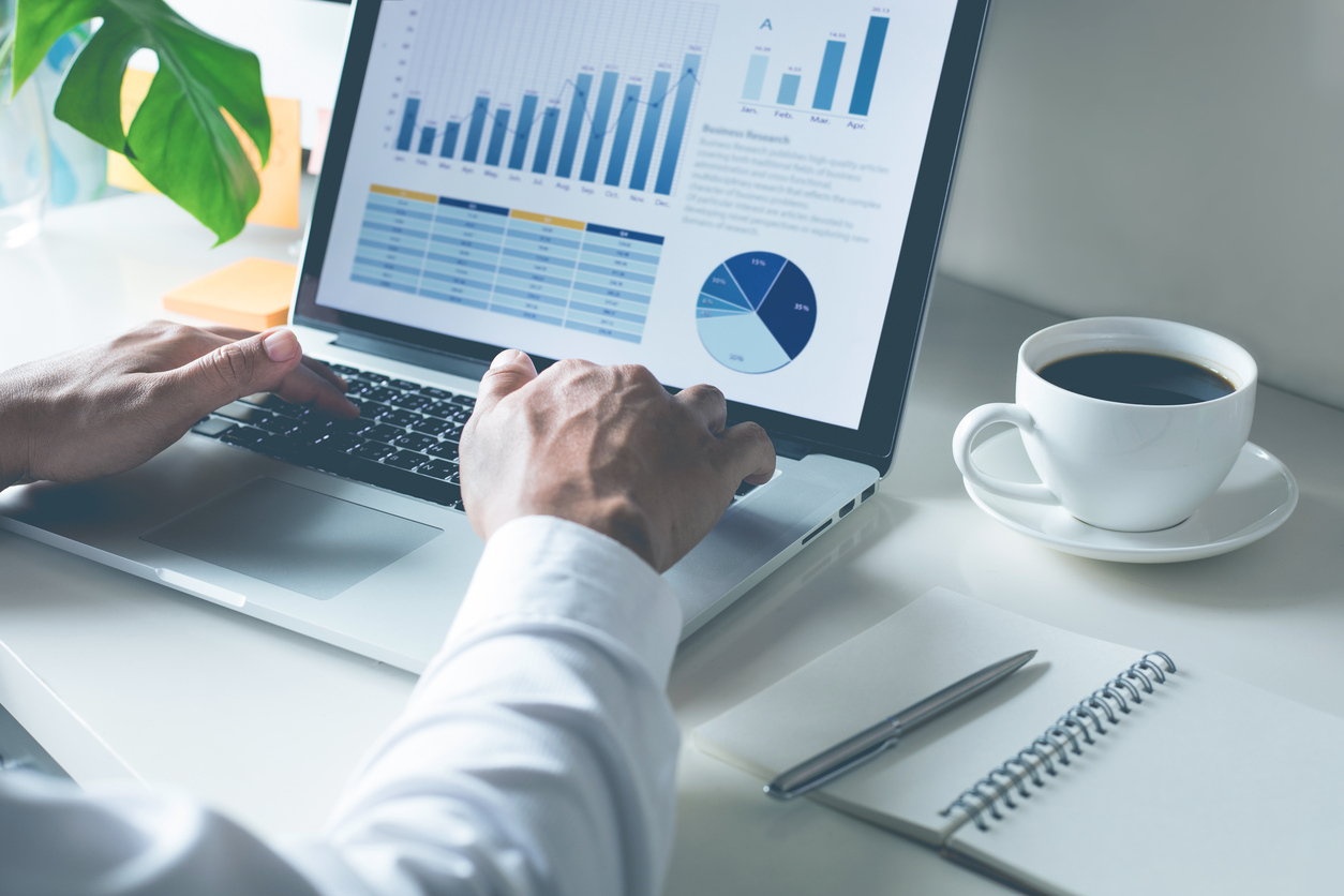 A person working at a laptop with graphs of data displayed on it. A plant, cup of coffee and notebook with pen are visible on the table around them.