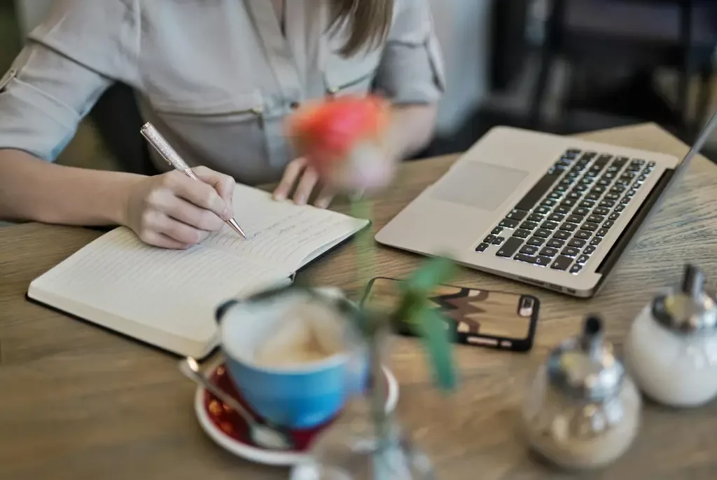A woman sitting at a table in a cafe, taking notes on a paper notepad in front of a laptop.